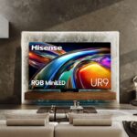 A large Hisense UR9 RGB MiniLED television is mounted on a textured, backlit marble wall above a floating wooden console in a modern, luxury living room. The TV screen displays a vibrant, abstract wave pattern of swirling blue, gold, and red lights, with the Hisense logo in the top left and "RGB MiniLED" and "UR9" text at the bottom. The room features a spacious beige sofa in the foreground, a dark lounge chair with an ottoman to the right, concrete-textured walls, and large floor-to-ceiling windows showing a city skyline view.