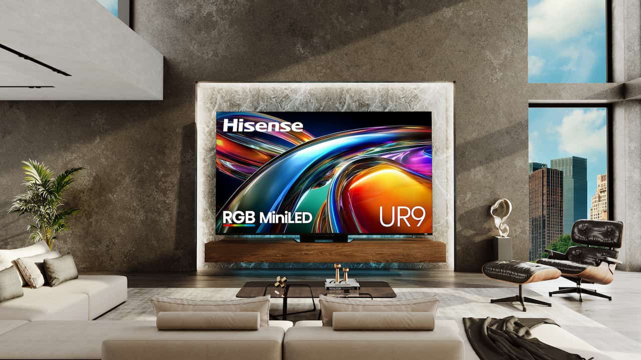 A large Hisense UR9 RGB MiniLED television is mounted on a textured, backlit marble wall above a floating wooden console in a modern, luxury living room. The TV screen displays a vibrant, abstract wave pattern of swirling blue, gold, and red lights, with the Hisense logo in the top left and "RGB MiniLED" and "UR9" text at the bottom. The room features a spacious beige sofa in the foreground, a dark lounge chair with an ottoman to the right, concrete-textured walls, and large floor-to-ceiling windows showing a city skyline view.