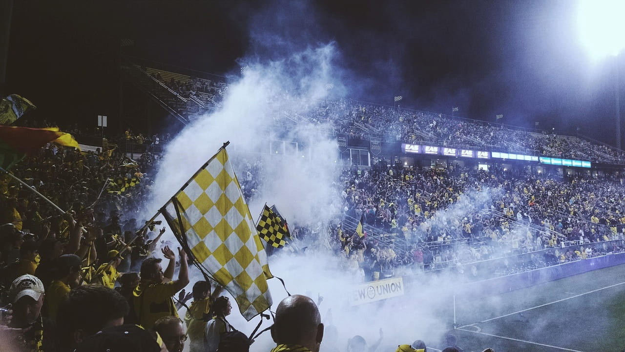 a packed sports stadium stand at night, filled with a large crowd of spectators dressed in yellow and black. Thick white smoke from flares drifts across the stands, partially obscuring the fans who are waving large checkered flags. A bright stadium floodlight illuminates the scene from the upper right, casting a strong glow over the noisy, high-energy environment.