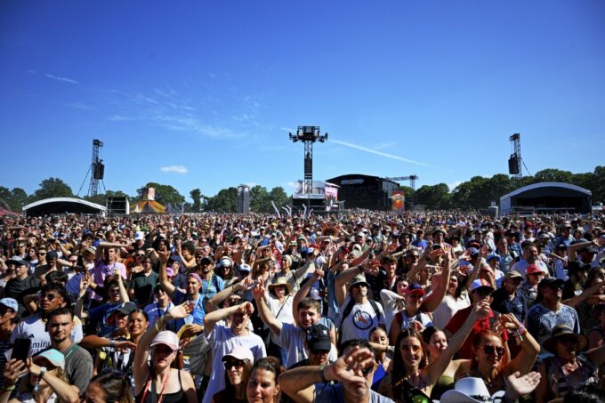 A vast crowd of festivalgoers standing outdoors in front of open-air stages under a clear blue sky, raising their hands and enjoying the event.