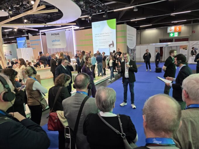 A colour photograph taken from behind a crowd of people looking towards a speaker at a busy exhibition booth. The attendees in the foreground are wearing large over-ear headphones to listen to the presentation. The speaker, a man in a dark blazer and jeans, stands on a blue carpeted floor holding a microphone and gesturing as he talks. In the background, large exhibition stands are visible