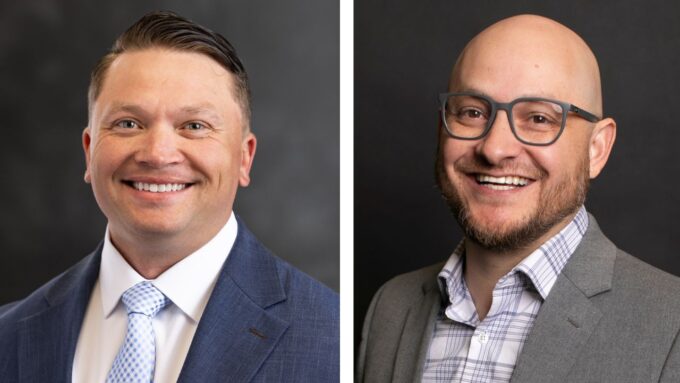 Side-by-side professional headshots against a dark background. On the left is a smiling man with dark hair wearing a blue suit jacket, white shirt, and patterned light blue tie. On the right is a smiling, bearded man with a shaved head and glasses, wearing a grey suit jacket over a patterned collared shirt.