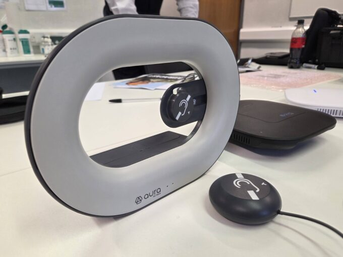 A close-up photograph on a table shows a distinctive, grey oval-shaped assistive listening device with "aura by Opus" branding. It has a central aperture containing a Telecoil/Auracast symbol (an ear with a 'T') on a swivel mount. In front of it rests a small, matching grey wired pendant receiver, also displaying the ear symbol. Other office equipment is visible but blurred in the background.