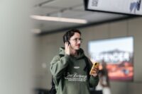 A male passenger with glasses and a green hoodie wears headphones and holds a smartphone inside an airport terminal, demonstrating how to use Auracast for announcements.
