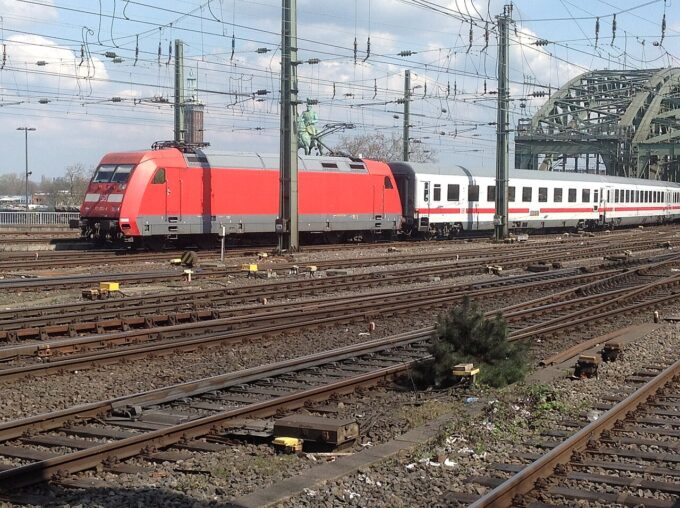 A red Deutsche Bahn Class 101 electric locomotive hauls white and red passenger carriages across a multi-track railway junction near the Hohenzollern Bridge in Cologne, with overhead electric lines criss-crossing above.