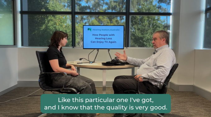A colour video screenshot showing Brooke Hudd and Brendan Lonergan sitting opposite each other in black office chairs at a round table in a bright room with large windows showing green trees outside. Brooke, on the left, is wearing a black top and dark trousers, looking towards Brendan. Brendan, on the right, wears a light-coloured patterned shirt and dark trousers, gesturing with his hand as he speaks. In the background on the table is a television screen displaying the text Hearing Matters Australia, How People with Hearing Loss Can Enjoy TV Again. A green caption box at the bottom of the screen reads: Like this particular one I've got, and I know that the quality is very good.