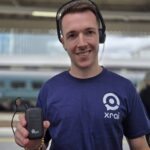 A man wearing a purple T-shirt with the XRAI logo holding the Ampetronic Auri system. Behind him is a train (it is blurred)