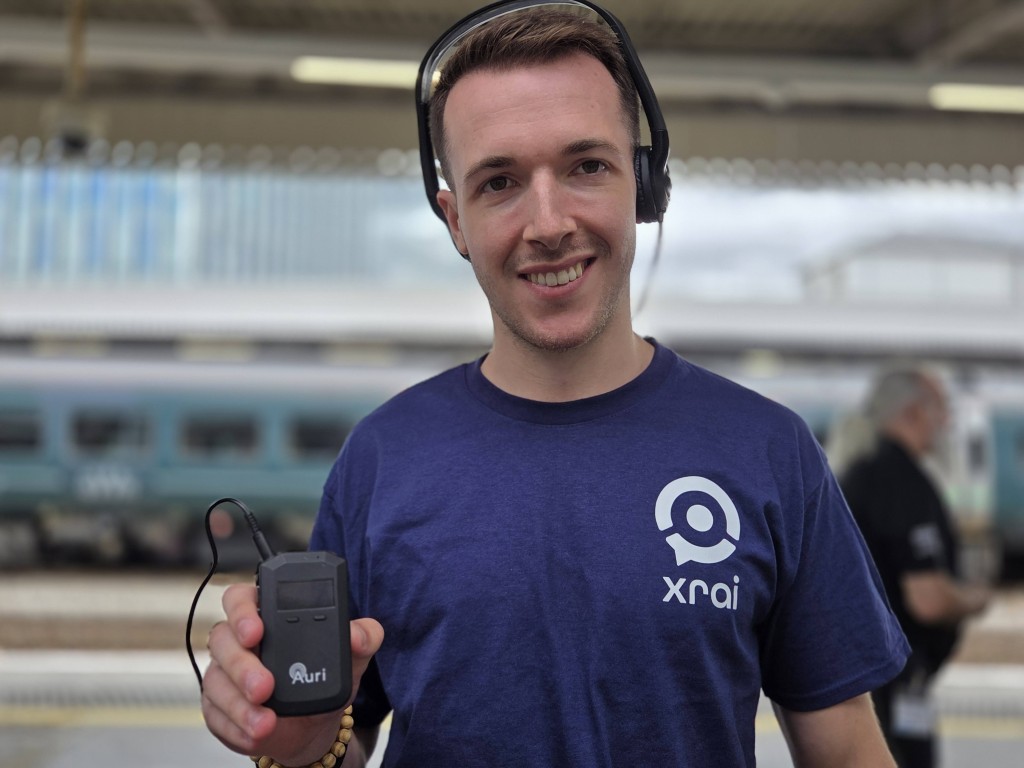 A man wearing a purple T-shirt with the XRAI logo holding the Ampetronic Auri system. Behind him is a train (it is blurred)