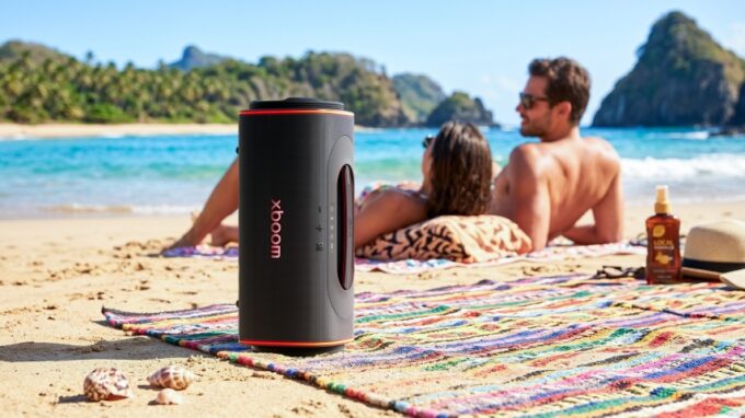 LG xboom Blast speaker standing vertically on a beach blanket with people relaxing near the ocean