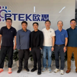 Senior leadership representatives from Optek, Audeara, and Eastech standing together in front of the Optek company logo on an office wall