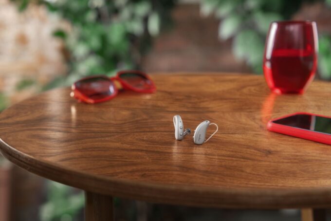 A pair of silver Signia Pure C&G MaX receiver-in-canal hearing aids standing on a round wooden table next to a smartphone