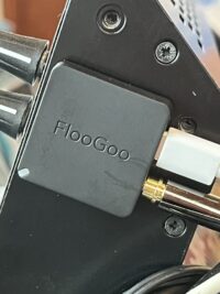 A close-up view of the back of a black sound mixing desk, where a small square FlooGoo FMA121 transmitter is affixed with BluTack. It has a white USB-C power cable and a gold-plated audio jack plugged into it
