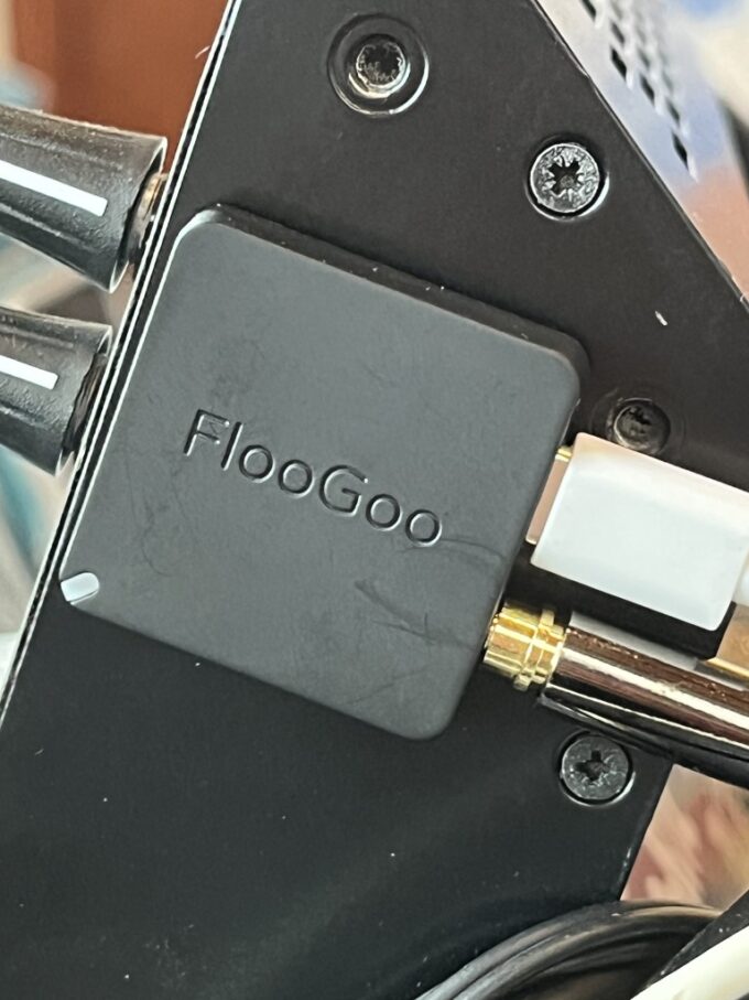 A close-up view of the back of a black sound mixing desk, where a small square FlooGoo FMA121 transmitter is affixed with BluTack. It has a white USB-C power cable and a gold-plated audio jack plugged into it