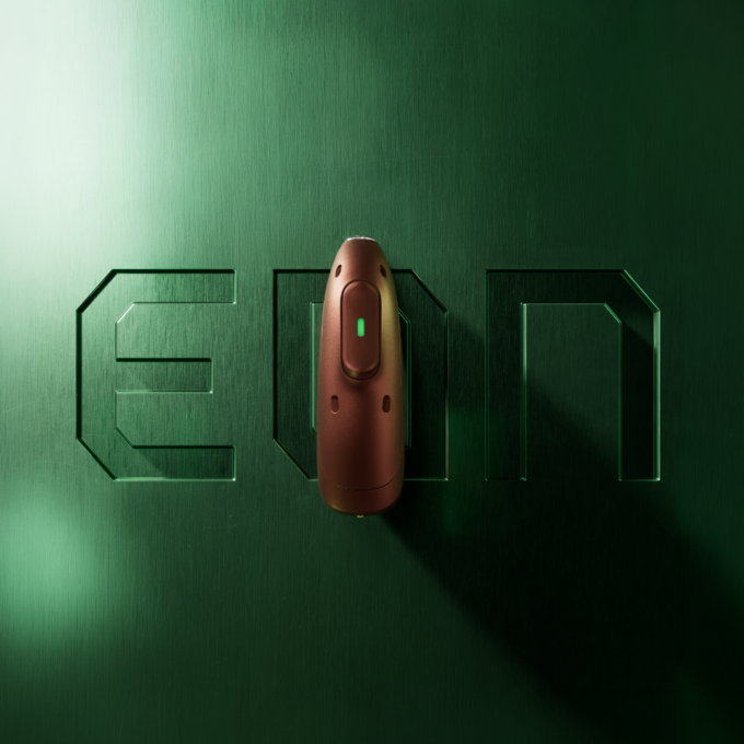 A sleek bronze hearing aid device with a glowing green indicator light resting vertically against an engraved green brushed metal surface