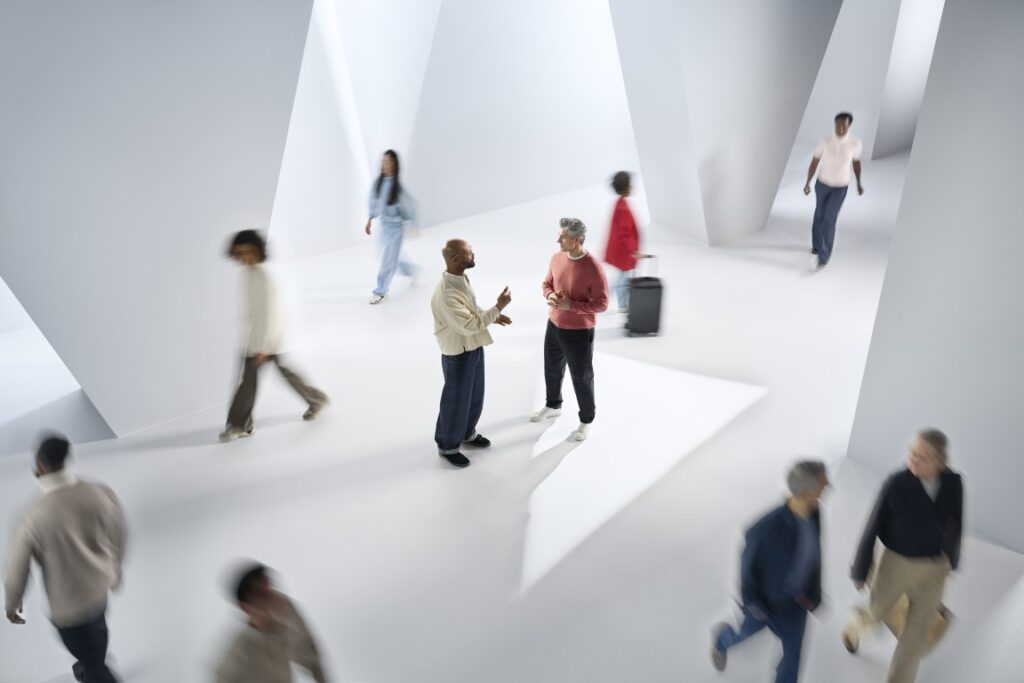 Two men stand in the centre of a large, modern hall with geometric walls, talking face-to-face and sharply in focus. Around them, multiple other people are walking past, blurred by motion, representing a busy background