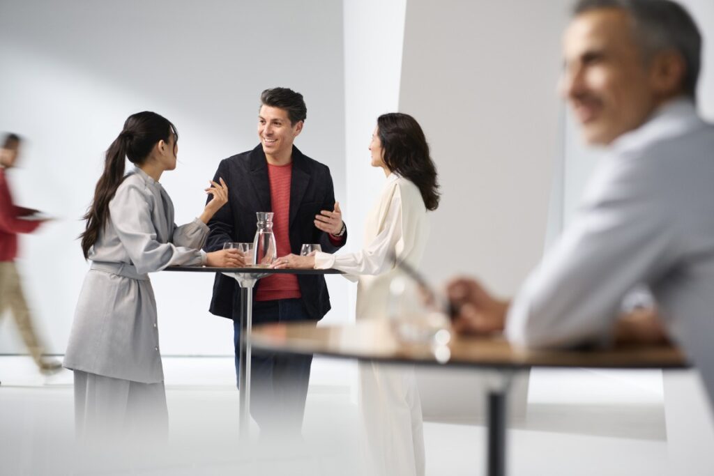 A sharp, focused group of three people (two women and one man) stands talking around a high-top table in a bright, modern indoor space. In the foreground, other people and tables are blurred. In the background, a person in a red top walks, also blurred by motion