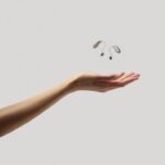 A person’s arm is extended, palm upward, with a pair of compact ReSound Sensia hearing aids floating in the air just above the hand. The hearing aids are light beige and dark grey. The background is a solid light grey.