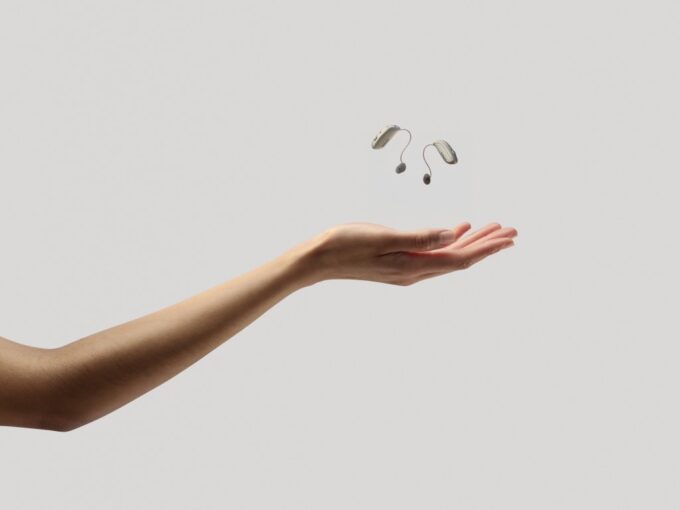 A person’s arm is extended, palm upward, with a pair of compact ReSound Sensia hearing aids floating in the air just above the hand. The hearing aids are light beige and dark grey. The background is a solid light grey.