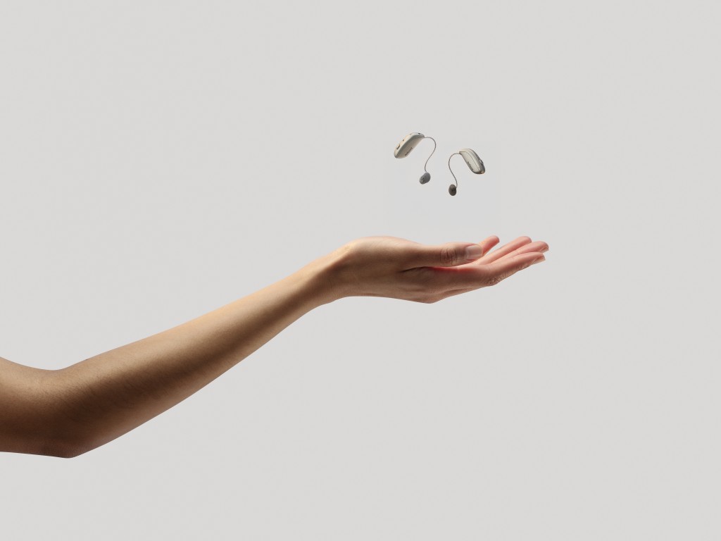 A person’s arm is extended, palm upward, with a pair of compact ReSound Sensia hearing aids floating in the air just above the hand. The hearing aids are light beige and dark grey. The background is a solid light grey.