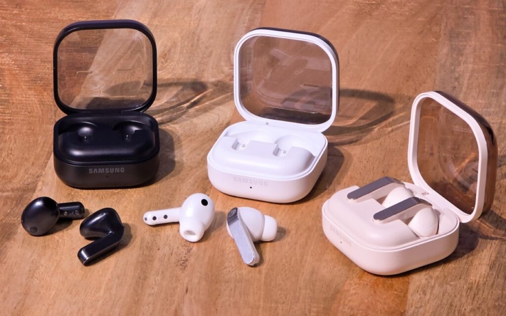 Three pairs of Samsung Galaxy Buds4 series wireless earbuds and open charging cases displayed in black, white, and cream color options on a wooden table
